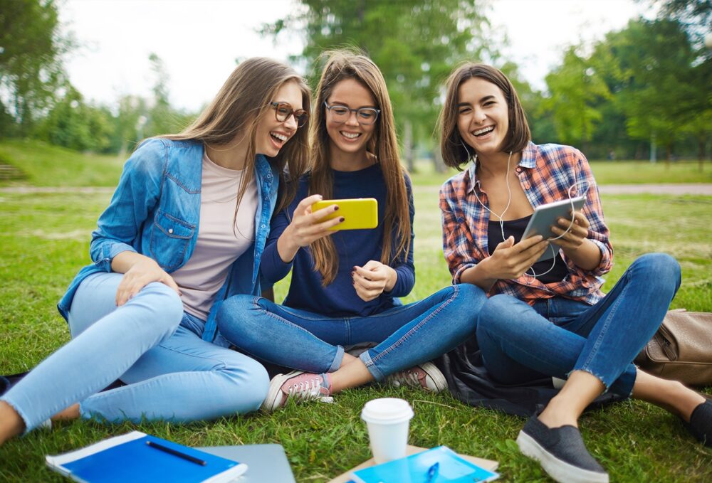 Three teenage girls compare smartphone photos in outdoor picnic.
